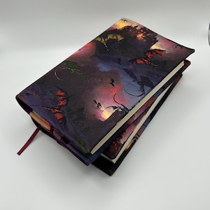 May include: Three hardcover books stacked, featuring a fantasy dragon-themed cover. The covers depict a dramatic scene with dragons flying against a castle and a fiery sky. A burgundy ribbon bookmark is visible, adding a touch of elegance.