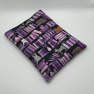Peut inclure: Une housse de coussin rectangulaire noire et violette avec un motif répété de livres et de chats. Les livres sont dans des tons violets et roses, et les chats sont dans diverses poses. La housse de coussin est en tissu.