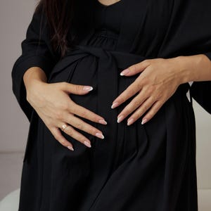 Puede incluir: Una bata de maternidad negra con un slip a juego debajo. La bata tiene un cinturón de atar y mangas largas. La modelo lleva la bata y se sujeta el vientre.