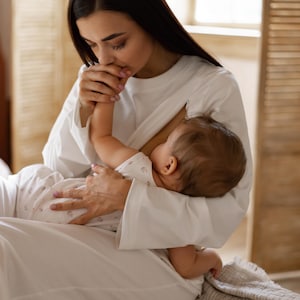 May include: A person wearing a white long-sleeve shirt and white pants, breastfeeding a baby. The baby is wearing a white onesie with a floral pattern. The scene is set indoors with a wooden screen and window in the background.
