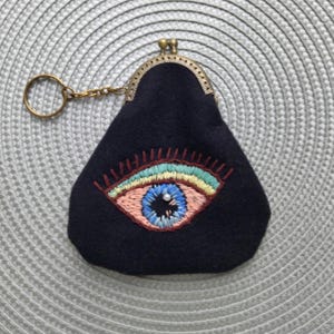 May include: A black coin purse with a brass clasp and key ring. The purse features an embroidered eye design with blue, pink, yellow, and green thread. The purse is made of a soft, dark fabric.