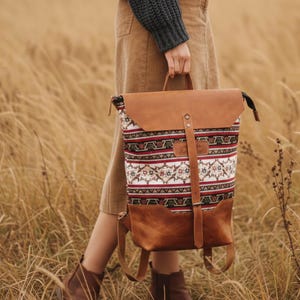 Könnte beinhalten: Ein brauner Lederrucksack mit einer gemusterten Stofffront. Der Stoff hat ein weißes, rotes und grünes Design. Der Rucksack hat eine obere Klappe und verstellbare Träger. Die Tasche wird in einem Feld mit hohem Gras gehalten.