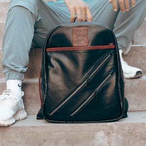 May include: Black backpack with red trim and two diagonal front pockets. The backpack has a top handle and a rectangular patch at the top. The backpack is sitting on concrete steps.