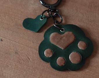 May include: Green leather keychain with a paw print design and a small heart charm. The paw print has brown leather accents.