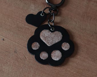 May include: Black leather keychain with a paw print design and a small heart charm. The paw print has a brown velvet heart in the center and brown velvet accents on the toes.