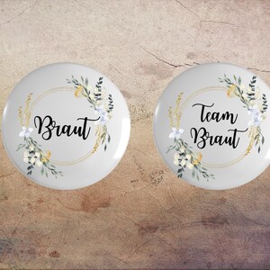 Könnte beinhalten: Zwei runde, weiße Knöpfe mit Blumenmuster und goldenen Akzenten. Der Text auf den Knöpfen lautet "Braut" und "Team Braut".
