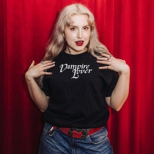 Puede incluir: Una camiseta negra de manga corta con el texto blanco Vampire Lover impreso en una fuente cursiva estilizada en el pecho. La camiseta se combina con unos vaqueros azules y un cinturón rojo con hebilla metálica.
