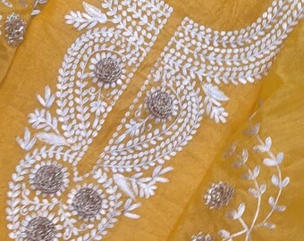 May include: Mustard yellow fabric with intricate white embroidery. The embroidery features floral and leaf patterns, with small, textured embellishments. The design is concentrated around the neckline and extends down the garment.