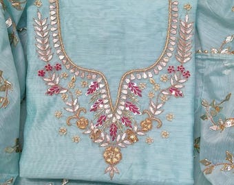 May include: A light blue embroidered salwar kameez set. The top features intricate gold and pink floral embroidery around the neckline. The matching dupatta and salwar have gold leaf accents. The fabric has a slight sheen.