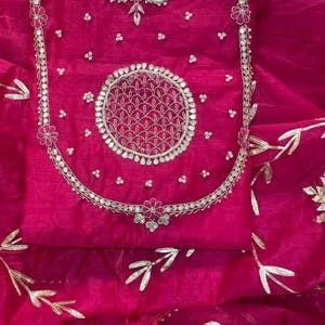 May include: A vibrant fuchsia-colored garment with intricate silver embroidery. The design features a central circular motif and floral accents. The fabric appears sheer, with delicate gold leaf patterns. This piece is likely a traditional ethnic wear.