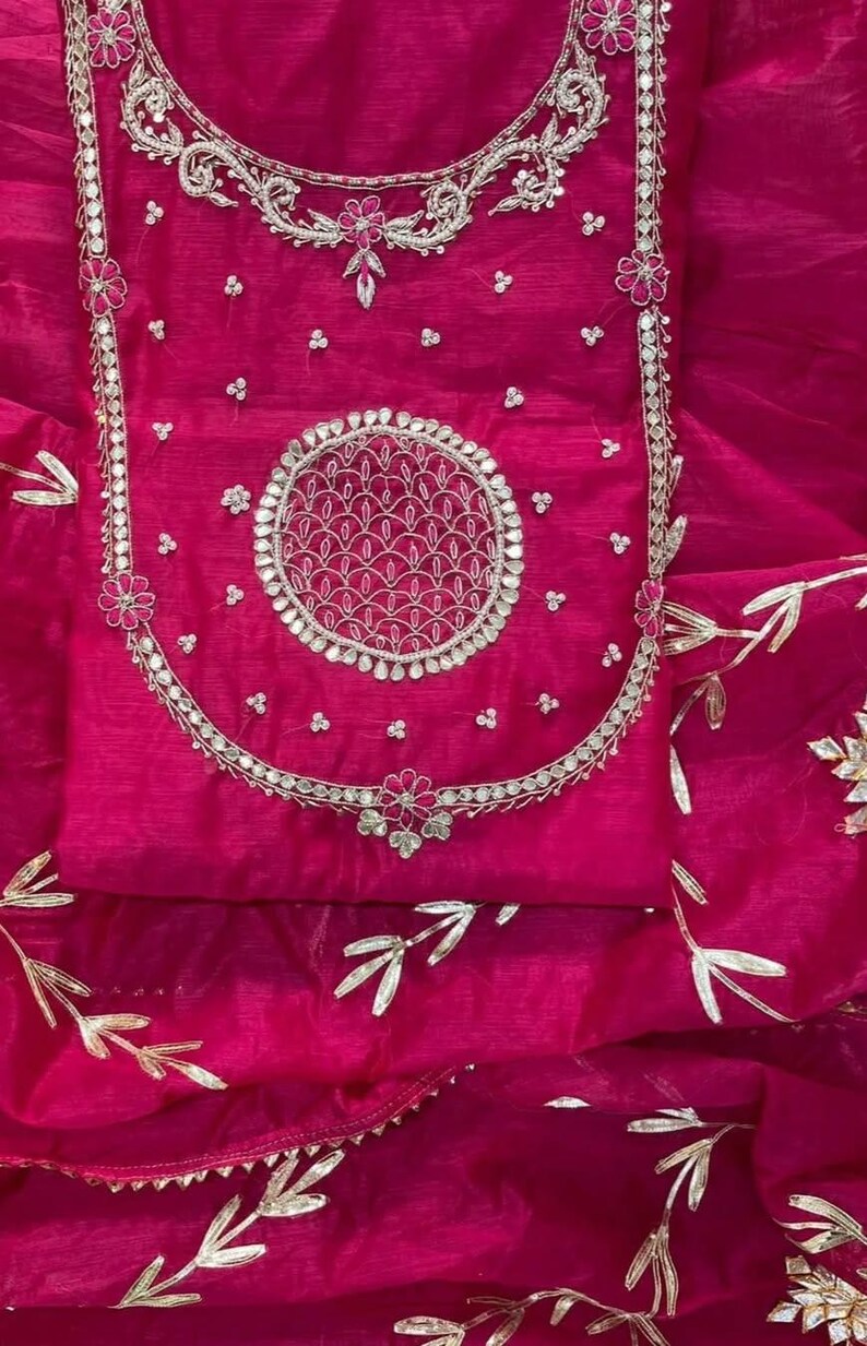 May include: A vibrant fuchsia-colored garment with intricate silver embroidery. The design features a central circular motif and floral accents. The fabric appears sheer, with delicate gold leaf patterns. This piece is likely a traditional ethnic wear.