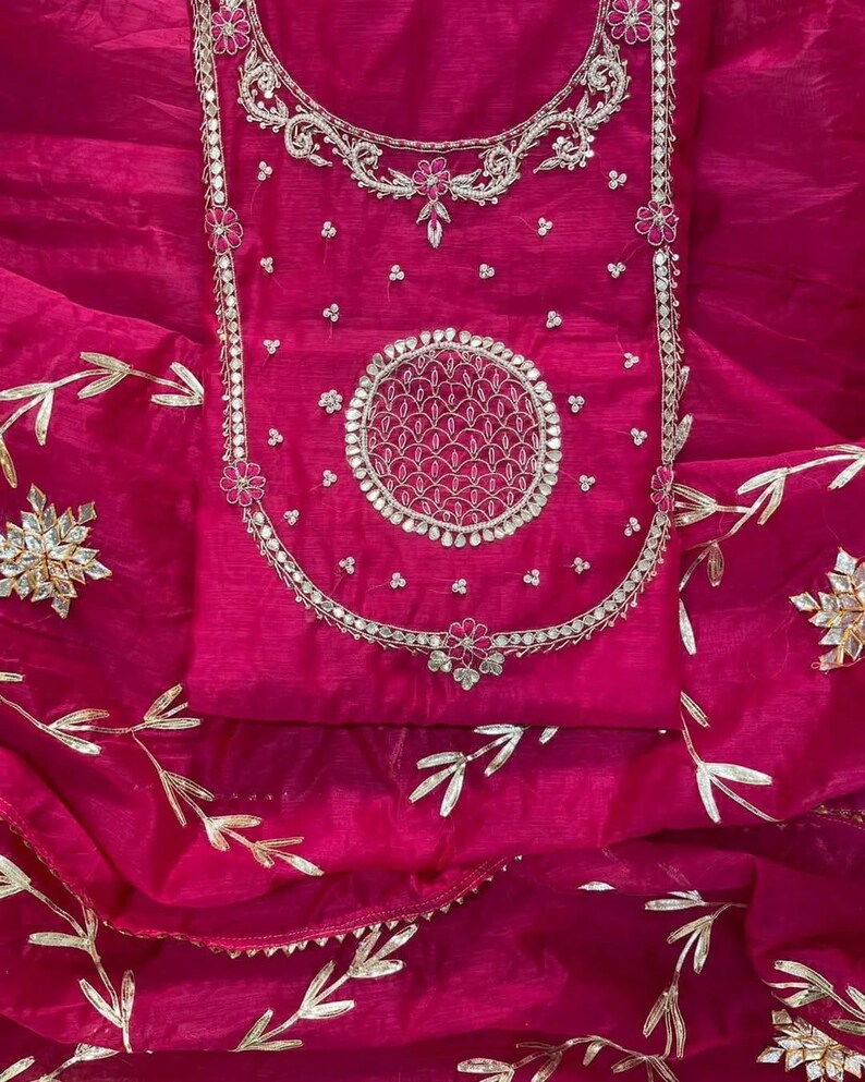 May include: A vibrant fuchsia-colored garment with intricate silver embroidery. The design features floral and circular patterns, with delicate gold leaf accents. The fabric has a sheer, flowing quality, suggesting a formal or festive attire.