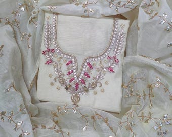 May include: Ivory-colored embroidered salwar kameez set. The top features a detailed floral design in silver and pink around the neckline. The matching dupatta and top are made of a sheer fabric with silver leaf accents.
