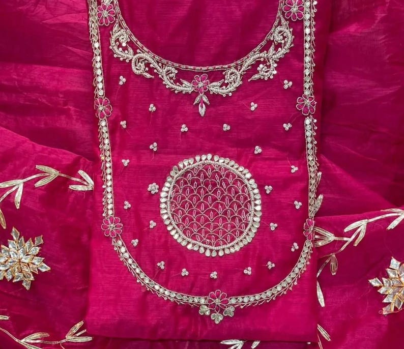 May include: A vibrant fuchsia-colored garment with intricate silver embroidery. The design features floral and geometric patterns, including a large circular motif. The fabric has a sheer, flowing quality, suggesting a formal or festive attire.