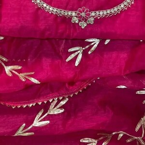 May include: A vibrant fuchsia-colored fabric with silver leaf-like embellishments. The fabric is draped, revealing a layered effect. A silver necklace with a floral design is visible at the top. The overall theme suggests a garment or textile.