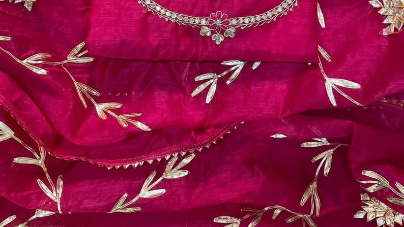 May include: A vibrant fuchsia-colored fabric with silver leaf-like embellishments. The fabric is draped, revealing a layered effect. A silver necklace with a floral design is visible at the top. The overall theme suggests a garment or textile.