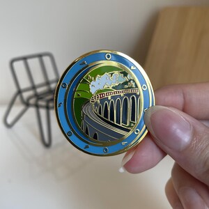 May include: A round, gold-toned pin with a blue border and a detailed illustration of a train crossing a bridge. The image features a train, green hills, and a blue sky. The pin is held by a person.