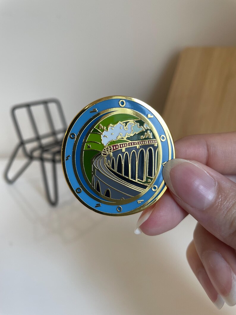 May include: A round, gold-toned pin with a blue border and a detailed illustration of a train crossing a bridge. The image features a train, green hills, and a blue sky. The pin is held by a person.