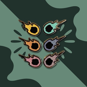 May include: Six comet-shaped enamel pins with glitter accents and gold-coloured metal borders. The pins feature a black centre and are arranged on a green background with a leaf-like pattern. Colours include yellow, teal, orange, purple, pink, and a second pink.