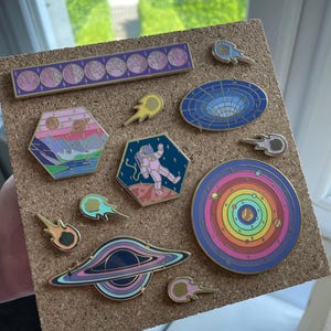 May include: A collection of enamel pins with space and astronomy themes. The pins feature designs of planets, astronauts, black holes, and comets in various colors, including pink, blue, and gold. The pins are displayed on a corkboard.