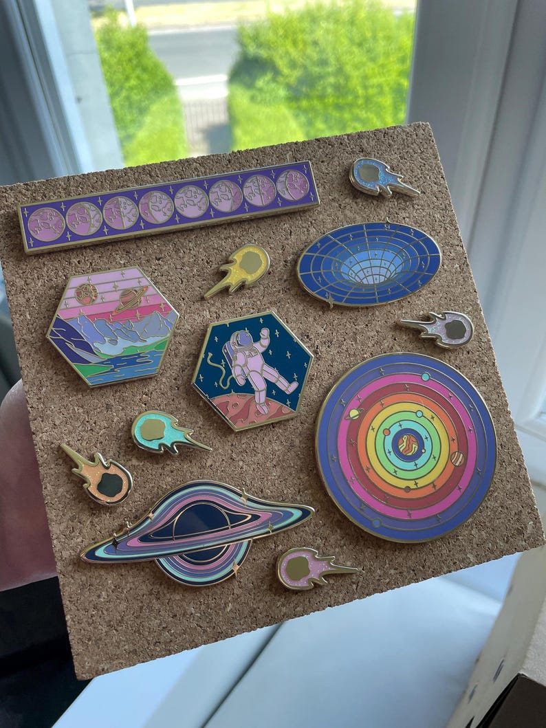 May include: A collection of enamel pins with space and astronomy themes. The pins feature designs of planets, astronauts, black holes, and comets in various colors, including pink, blue, and gold. The pins are displayed on a corkboard.
