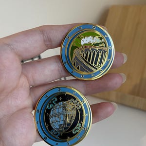 May include: Two round enamel pins with gold trim. One pin features a train on a bridge, the other a building with the letters "SR". Both pins have a blue border and black and gold accents.