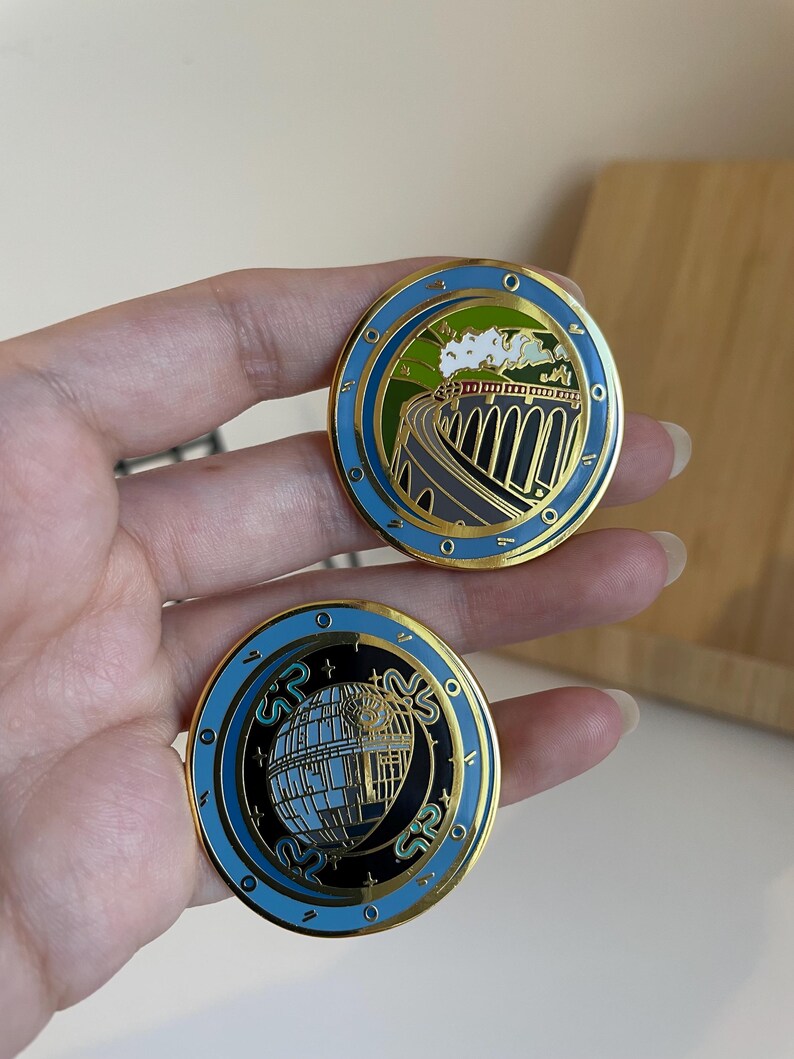 May include: Two round enamel pins with gold trim. One pin features a train on a bridge, the other a building with the letters "SR". Both pins have a blue border and black and gold accents.