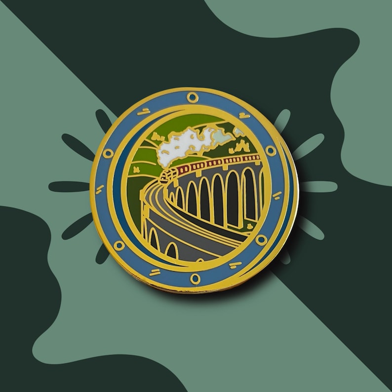 May include: A round enamel pin featuring a train traveling over a stone bridge. The pin has a blue border with gold accents and a green and yellow interior. The train emits white smoke against a blue sky.