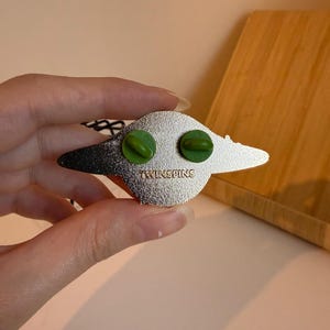 May include: A silver-colored enamel pin with a stylized design. The pin features two green pin backs and the text "TWINSPINS" stamped on the back. The pin has a black and silver color scheme.