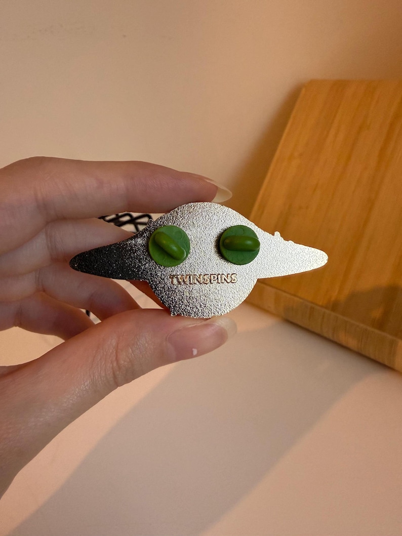 May include: A silver-colored enamel pin with a stylized design. The pin features two green pin backs and the text "TWINSPINS" stamped on the back. The pin has a black and silver color scheme.