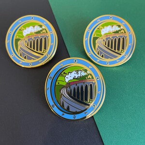 May include: Three circular enamel pins featuring a train crossing a stone viaduct. The pins have a gold border, a blue outer ring, and a green and blue background. The train is red and white, and the sky is white.