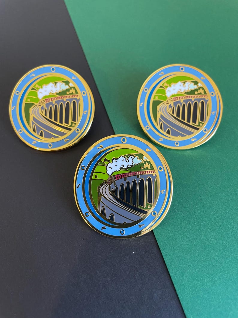 May include: Three circular enamel pins featuring a train crossing a stone viaduct. The pins have a gold border, a blue outer ring, and a green and blue background. The train is red and white, and the sky is white.