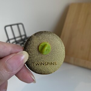 May include: A gold-colored round pin with a textured surface. The pin has a green clasp and the word "TWINSPINS" is printed on the surface. The pin is held by a person.