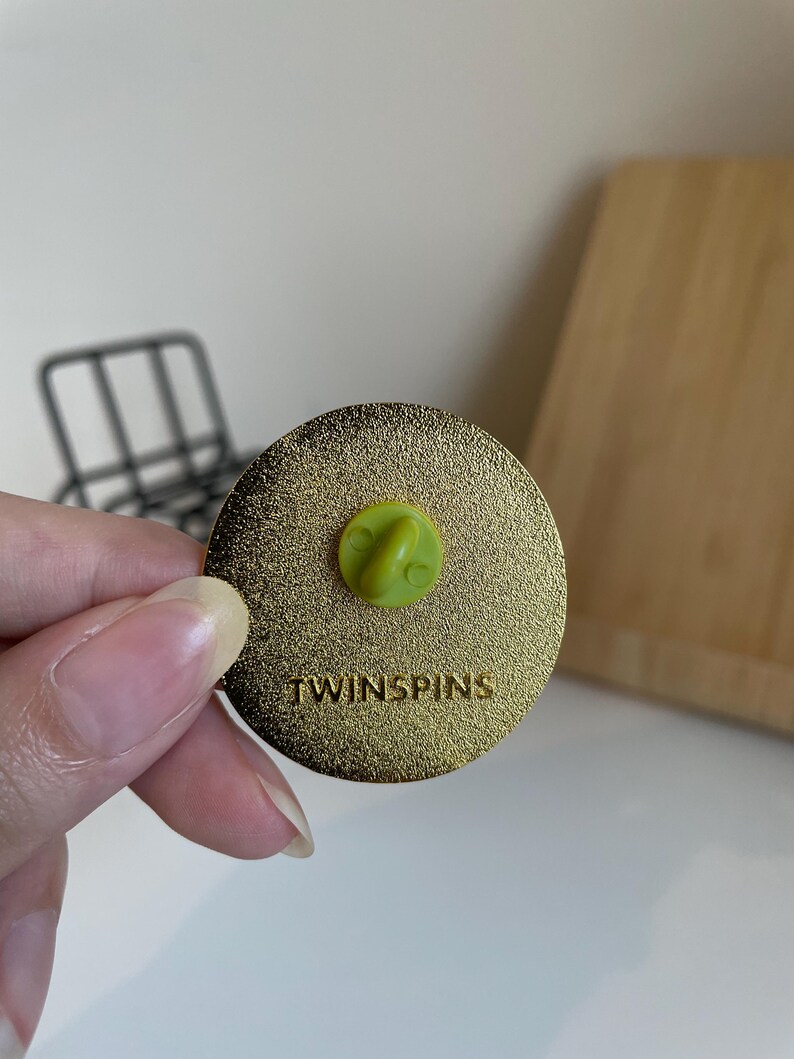 May include: A gold-colored round pin with a textured surface. The pin has a green clasp and the word "TWINSPINS" is printed on the surface. The pin is held by a person.