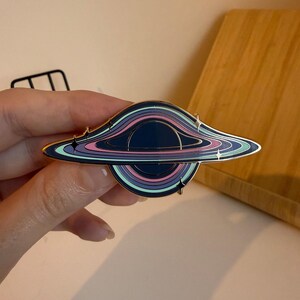 May include: A black hole enamel pin with a gold border. The pin features concentric rings of color, including blue, purple, pink, and teal. Small star-shaped accents are placed around the edges of the black hole design.