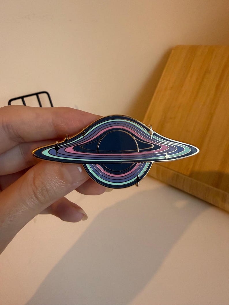 May include: A black hole enamel pin with a gold border. The pin features concentric rings of color, including blue, purple, pink, and teal. Small star-shaped accents are placed around the edges of the black hole design.