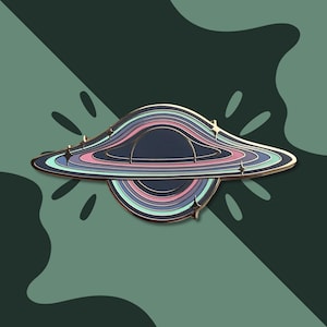 May include: A black hole enamel pin with a gold border. The pin features a dark blue center with concentric rings in shades of pink, blue, and teal. Small star-shaped accents are placed around the outer edge. The background is a dark green with a leaf-like pattern.