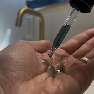 May include: A hand with a clear glass dropper bottle with a black cap. The bottle is dispensing a dark green liquid onto the palm of the hand.