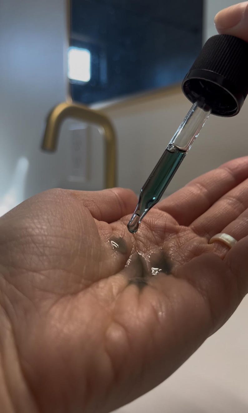 May include: A hand with a clear glass dropper bottle with a black cap. The bottle is dispensing a dark green liquid onto the palm of the hand.