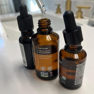 May include: Three amber glass bottles with black dropper tops, filled with a clear liquid. The centre bottle has a white label with the text "HOW TO USE Apply to skin & hair as needed. Appliquer au besoin sur la peau et les cheveux" and several small green and pink certification logos. The bottle on the right has a white label with the text "30 mL" and a black label with text in a foreign language.