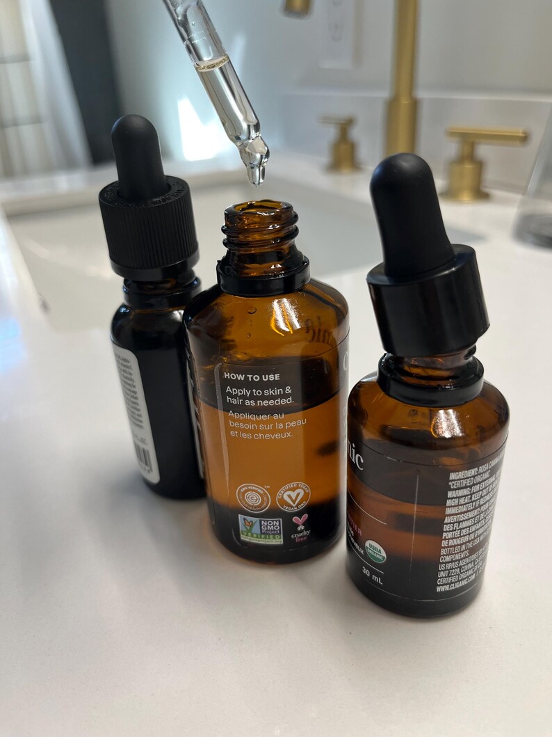 May include: Three amber glass bottles with black dropper tops, filled with a clear liquid. The centre bottle has a white label with the text "HOW TO USE Apply to skin & hair as needed. Appliquer au besoin sur la peau et les cheveux" and several small green and pink certification logos. The bottle on the right has a white label with the text "30 mL" and a black label with text in a foreign language.
