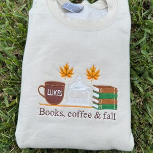 May include: Beige sweatshirt with embroidered design. Features a brown coffee mug with "Lukes" text, a white gazebo, and stacked books. Two orange leaves are above the gazebo. The text "Books, coffee & fall" is below the design.