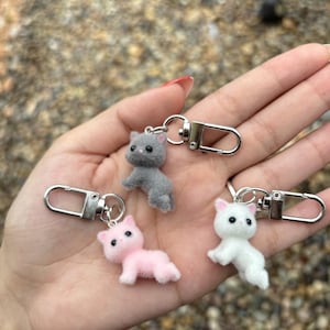 May include: Three plush cat keychains with silver metal clasps. The cats are gray, pink, and white.