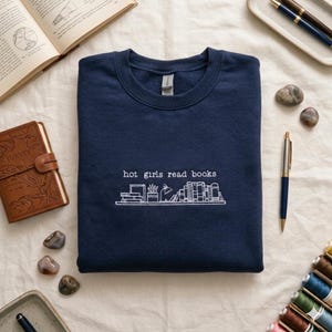 May include: A navy blue crewneck sweatshirt featuring white embroidery on the chest. The design includes the text "hot girls read books" above a line drawing of a bookshelf containing various books, a potted plant, and a small desk lamp.