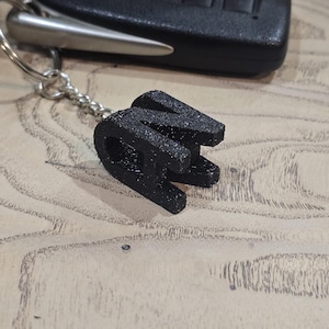 May include: A black, 3D-printed letter "W" keychain with a silver keyring. The keychain is next to a black car key fob on a light wood-grain surface.