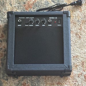 Könnte beinhalten: Schwarzer Gitarrenverstärker mit der Aufschrift "Guitar Amplifier" und "OYD-15". Er verfügt über Knöpfe für Drive Volume, Höhen und Bässe sowie Eingangs- und Telefonanschlüsse. Ein Netzkabel ragt oben heraus.