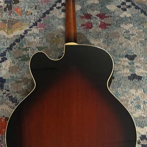 May include: Acoustic guitar with a sunburst finish, showcasing a gradient from dark brown to reddish-brown. The guitar's back and neck are visible, with silver tuning pegs and a cream-colored binding around the body. The instrument rests on a patterned rug.