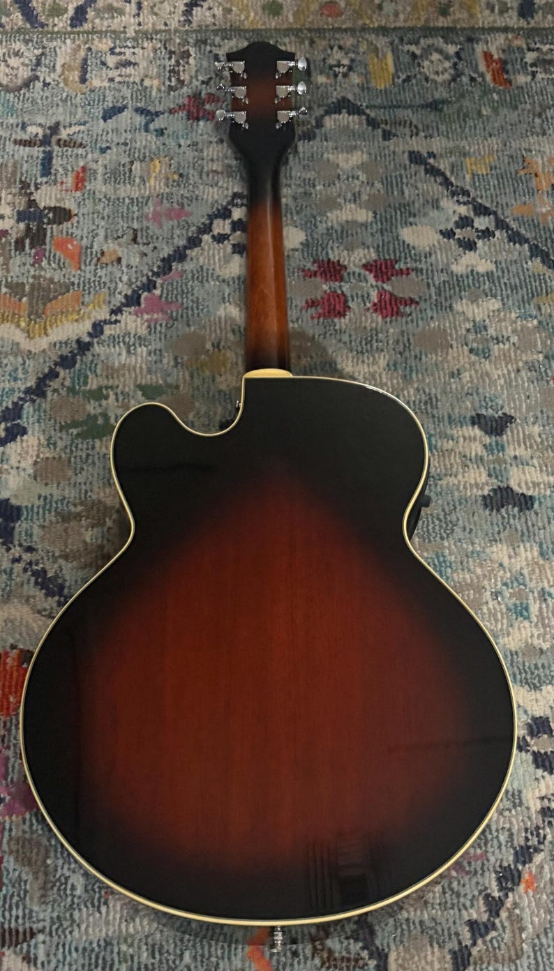 May include: Acoustic guitar with a sunburst finish, showcasing a gradient from dark brown to reddish-brown. The guitar's back and neck are visible, with silver tuning pegs and a cream-colored binding around the body. The instrument rests on a patterned rug.