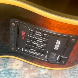 May include: Close-up of a Gretsch acoustic guitar with a black control panel. The panel has the word "PREFIX" in red, along with controls for bass, contour, treble, and volume. The guitar's body is brown and black.