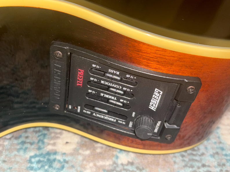 May include: Close-up of a Gretsch acoustic guitar with a black control panel. The panel has the word "PREFIX" in red, along with controls for bass, contour, treble, and volume. The guitar's body is brown and black.